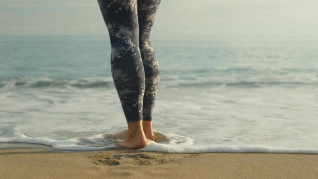 The legs of a girl in leggings, on a sandy sea beach, foam from the water touches her legs. Close-up, slow motion