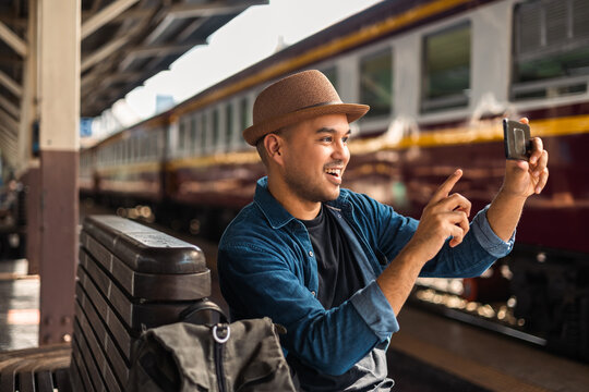 Freedom traveler young asian man at train station taking photo by smartphone. Happy tourist travel by train on vacation time holiday weekend trip. Backpacker arrival at platform railway.