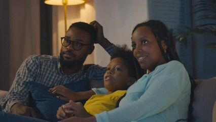 Cheerful African-American family with kid resting on couch talking and watching tv in evening - Powered by Adobe