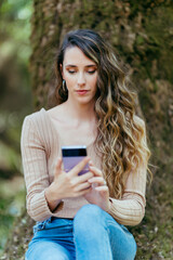 Naklejka na ściany i meble Chica joven con pelo largo ondulado en sendero verde con smartphone interactuando y haciendo fotos 