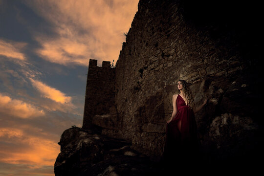 Chica joven con vestido rojo en zona boscosa y castillo medieval posando