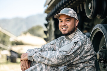 Fototapeta premium Asian man special forces soldier against on the field Mission. Commander Army soldier military defender of the nation in uniform sitting near battle tank while state of war.