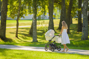 Young adult mother in dress pushing white baby stroller and walking on sidewalk at town park in warm sunny summer day. Spending time with infant and breathing fresh air. Enjoying stroll. Back view. - Powered by Adobe