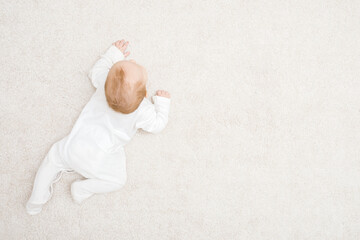 Baby in white bodysuit crawling on knee and arms on light beige home carpet background. Top down...