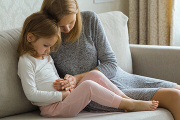 Child sitting on sofa and suffering from stomach ache at home room. Young adult mother hand touching daughter painful belly and trying calm down. - obrazy, fototapety, plakaty