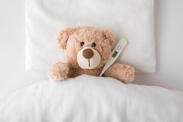 Teddy bear with digital thermometer lying down on pillow and sheet under blanket in white bed....