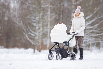 Fototapeta na wymiar Smiling young adult mother pushing white baby stroller and walking on snow covered sidewalk at park in cold winter day. Spending time with newborn and breathing fresh air. Enjoying peaceful stroll.