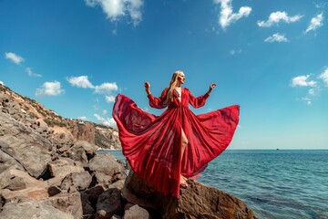 Red dress sea woman. A blonde with flowing hair in a long flowing red dress stands on a rock near the sea. Travel concept, photo session at sea