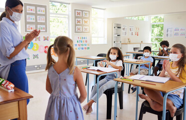 Covid, education and learning with a teacher wearing a mask and clapping for a student after her oral with classmates in class during school. Young girl talking or sharing her answer in a classroom - Powered by Adobe