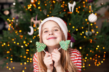 Close up of smiling girl wearing striped pajamas and Santa Claus hat with two lollipops in the...