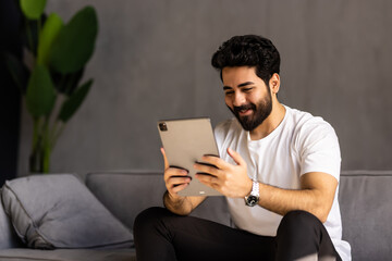 Young happy man at home reading on tablet on beige couch in light livingroom