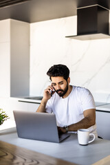 Smiling young arab man freelancer talking on mobile phone and taking notes, working from home during isolation, copy space. Happy guy having conversation on phone, using laptop at kitchen