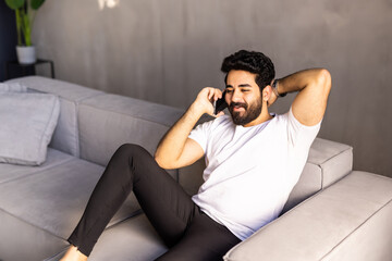 Obraz premium Cheerful arab man with long hair and glasses relaxing on couch at home, having phone talk with friends, looking at copy space and smiling, wearing casual outfit, resting at weekend