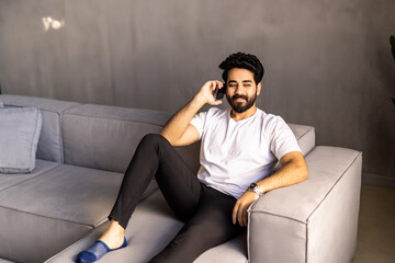 Fototapeta premium Cheerful arab man with long hair and glasses relaxing on couch at home, having phone talk with friends, looking at copy space and smiling, wearing casual outfit, resting at weekend