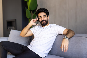 Happy emotional arab guy having phone conversation at home, got good news, sitting on sofa in living room.