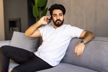 Fototapeta premium Happy emotional arab guy having phone conversation at home, got good news, sitting on sofa in living room.