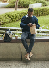 Business man eating food for lunch on his break outdoors at a park opening a brown paper bag. Hungry, happy black male time to eat and drink outside in the city. Enjoying a takeaway or takeout meal