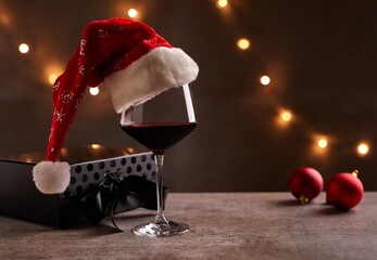 Glass of red wine with Santa Claus hat, bronze gift box and Christmas decorations on table. Christmas lights in the black background.