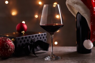 Glass of red wine with Santa Claus hat, black gift box and Christmas decorations on table. Christmas lights in the background.