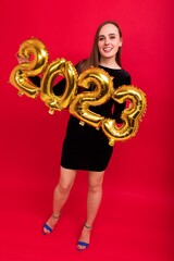 A young woman in a black evening dress and high-heeled shoes holds balloons in the shape of the numbers of the 2023 new year on a red background