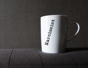 White coffee mug on dark gray background with printed text NARCISSIST , self-centered inflated...