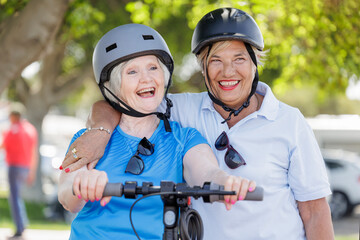 coppia di anziane con caschetto di protezione si abbraccia felice in una pista ciclabile con vicino un monopattino elettrico 