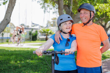 coppia di anziani coniugi vestiti sportivi e con caschetto dio protezione in testa tengono vicino  un monopattino elettrico  mentre stazionano in una pista ciclabile.
