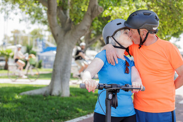 coppia di anziani coniugi vestiti sportivi e con caschetto dio protezione in testa tengono un monopattino elettrico  - I due si baciano appassionatamente in una pista ciclabile