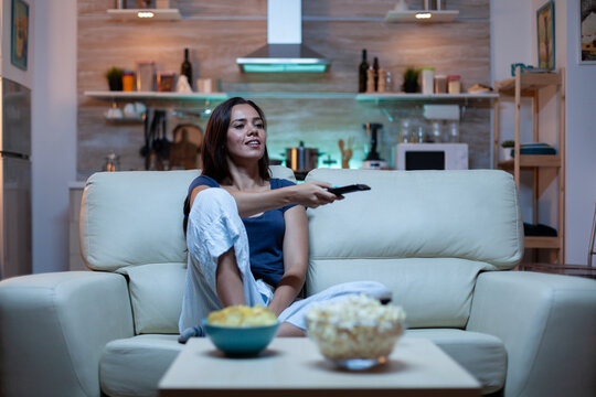 Caucasian woman sitting on couch and watching tv relaxing after work. Excited amused home alone lady in pijamas resting with snacks and juice sitting on comfortable sofa in open space living room.