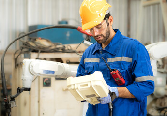 Male industrial engineer using remote control board to check robotic welder operation in modern automation factory. Technician worker monitoring robot controller system for automated steel welding.