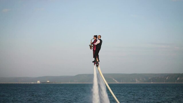Show over the water on a flyboard. A woman in a red dress plays the saxophone, a man flies with her together.