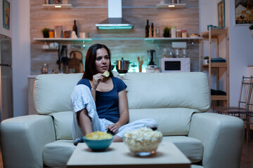 Happy woman eating popcorn on sofa and watching tv in living room at home. Excited, amused, lonely lady enjoying the evening sitting on comfortable couch dressed in pajamas in front of television.
