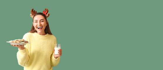 Happy young woman in deer horns holding plate with gingerbread cookies and glass of milk on green...