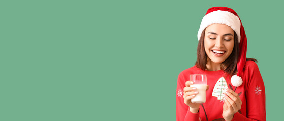 Happy young woman in Santa hat, with gingerbread cookie and glass of milk on green background with space for text