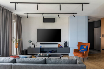 Interior design of living room interior with big tv, gray sofa, orange armchair, patterned pillow, gray curtain, wooden floor, vase with dried flowers and personal accessories. Home decor. Template.