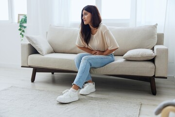 Woman sitting on sofa at home back and stomach pain during menstruation, women's day, women's health problems, suspected appendicitis