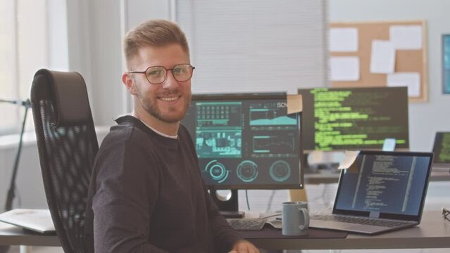 Medium portrait of Caucasian male programmer in eyeglasses turning to camera and starting smiling, sitting by office desk with program codes and schemes on computer and laptop screens
