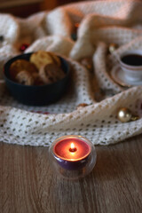 Cups of tea or coffee, bowl of cookies, soft blanket, lit candles, books and various Christmas decorations on the table. Selective focus.