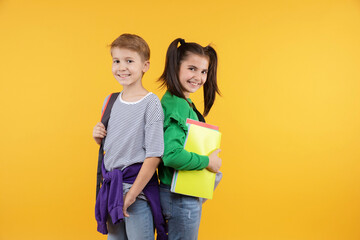 Smiling schoolkids in trendy outfits posing on yellow background. Positive kids holding backpacks and books, smiling, looking to camera. Studio shot. Back to school.