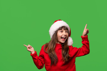 A cheerful smiling girl in a Santa hat shows with her index fingers discounts, promotions, gifts. Emotional portrait isolated on green background. New Year and Christmas concept.