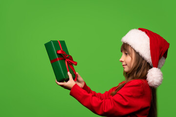 Cute little Santa girl in a hat holds a New Year's gift in her hands and looks at him. Happy New Year. Isolated on green background
