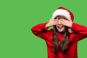 The girl in the Santa hat covered her eyes with her hands and stuck out her tongue. Emotional portrait of a child isolated on a green background. New Year mood