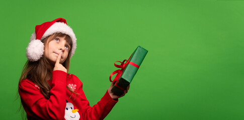 A cute little Santa girl in a hat holds a New Year's gift in her hands, looks up dreamily and tries to guess the surprise in the box. Merry Christmas and Happy New Year. Isolated on green background