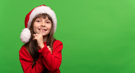 Don't give a surprise. Cute smiling little Santa girl in a hat put her finger to her lips. Silence sign. New Year and Christmas concept. Time to give gifts