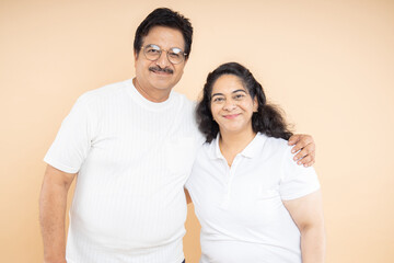 Portrait of happy Indian senior couple wearing plane white t-shirt standing isolated on beige studio background. Asian mature man and woman looking at camera
