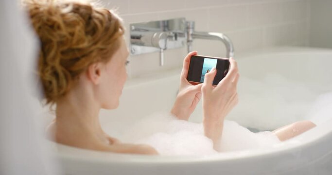 Young woman on her phone going through and zooming in on photos while she is relaxing in a bubble bath. Female enjoying her self care bathtub with hot water in the bathroom.