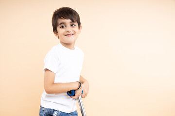 Closeup of smiling indian boy kid wearing white t-shirt rides holding scooter or bicycle handle on beige background with copy space. Studio shot, Childhood and active leisure concept.