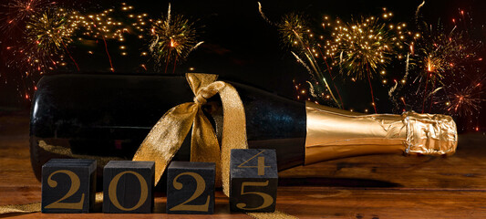 New Year 2024 / 2025 Sylvester New Year's Eve celebration holiday greeting card banner - Black cubes with year on sparkling wine or champagne bottle on table and firework in the background