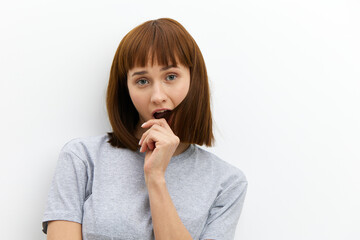 a sad, upset woman stands on a white background in a gray T-shirt and brings a lock of hair to her mouth, looking at the camera with an annoyed look