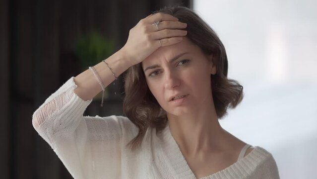A young woman at home touches her forehead with her hand to check her body temperature, suffering from a headache. Migraine attack in an adult. The first symptoms of a respiratory viral disease.